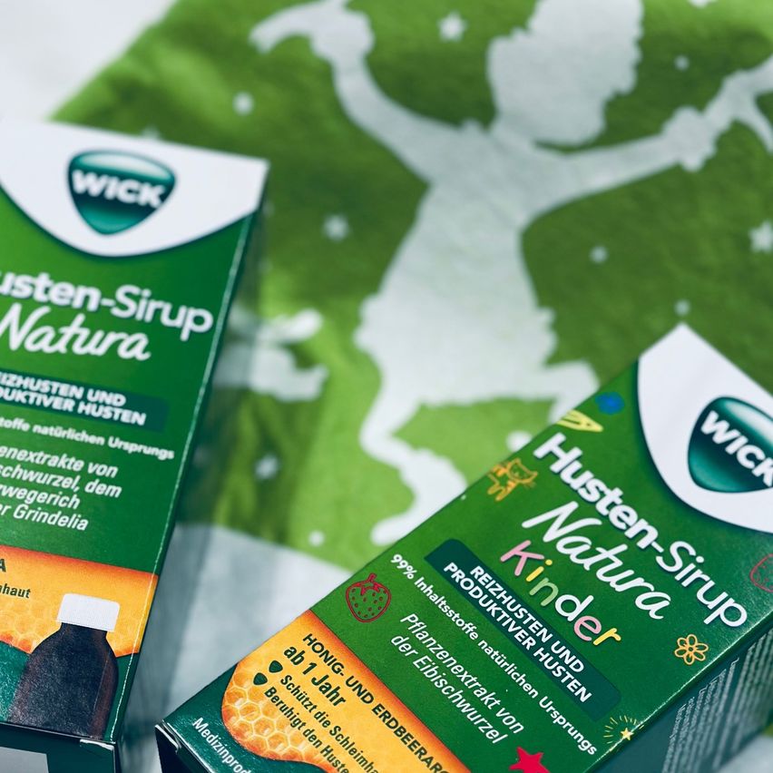 Zwei Schachteln Wick Husten-Sirup Natura stehen auf einem grünen Hintergrund. Eine Schachtel ist für Kinder und die andere für Erwachsene. Beide haben Etiketten, die angeben, dass sie natürliche Inhaltsstoffe enthalten.