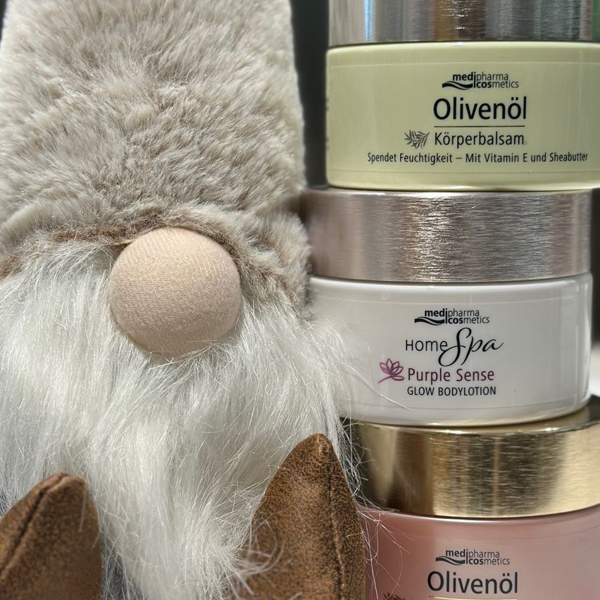 Drei Kosmetikflaschen und eine Gnomfigur sind zu sehen. Die oberste Flasche ist mit 'Olivenoel Korperbalsam' und zusätzlichen Text beschriftet. Darunter befindet sich eine weiße Flasche mit der Aufschrift 'Home Spa Purple Sense Glow Bodylotion.' Darunter ist eine rosa Flasche mit der Aufschrift 'Olivenoel.'