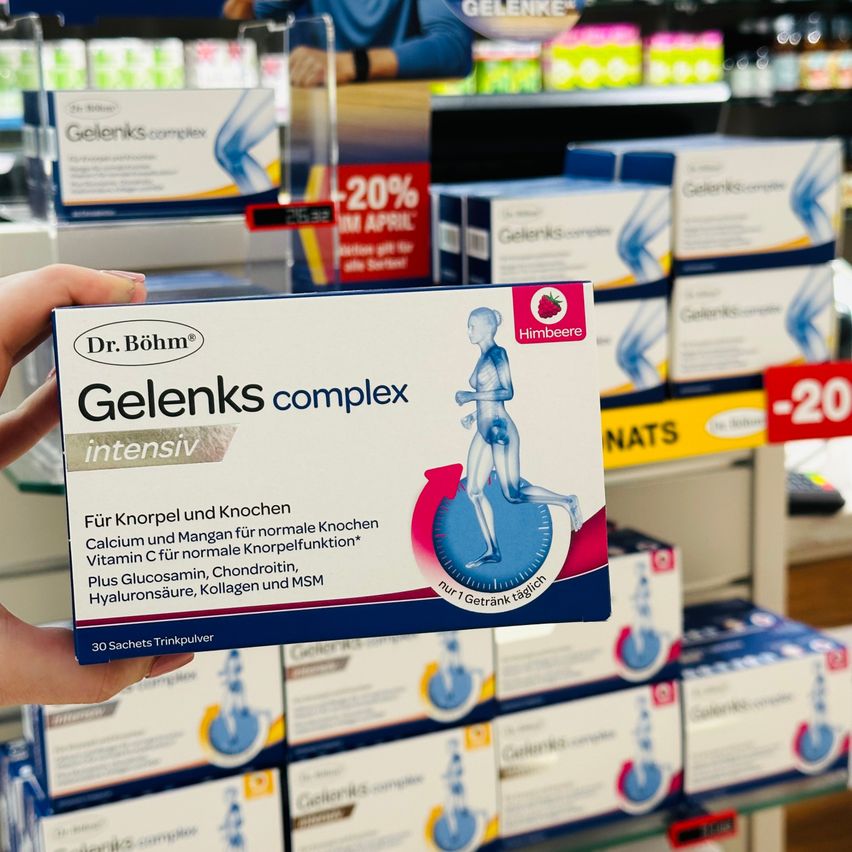 A hand holds a box of Gelenks complex, a product with a blue and white label featuring a silhouette of a person exercising. The box is located in a pharmacy with shelves of other health products in the background.