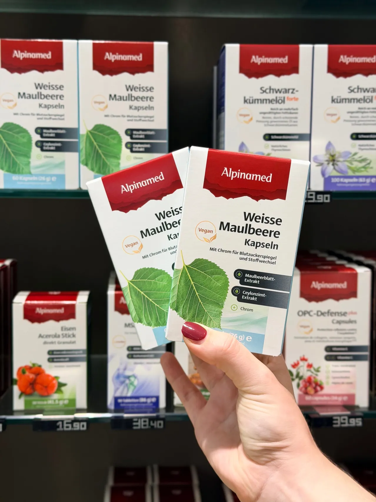 Eine Hand hält zwei Schachteln Alpinemed weiße Maulbeerkapseln in einem Geschäft, mit anderen Produkten im Hintergrund.