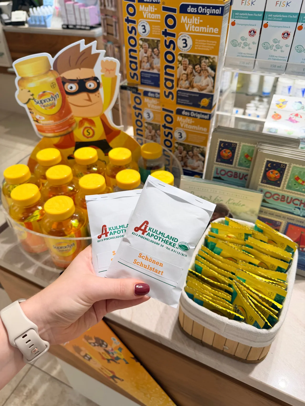 Eine Person hält zwei Packungen Kulmland Apothke Schoenen Schulstart vor einem Display mit Flaschen von Supredyn und anderen Produkten.