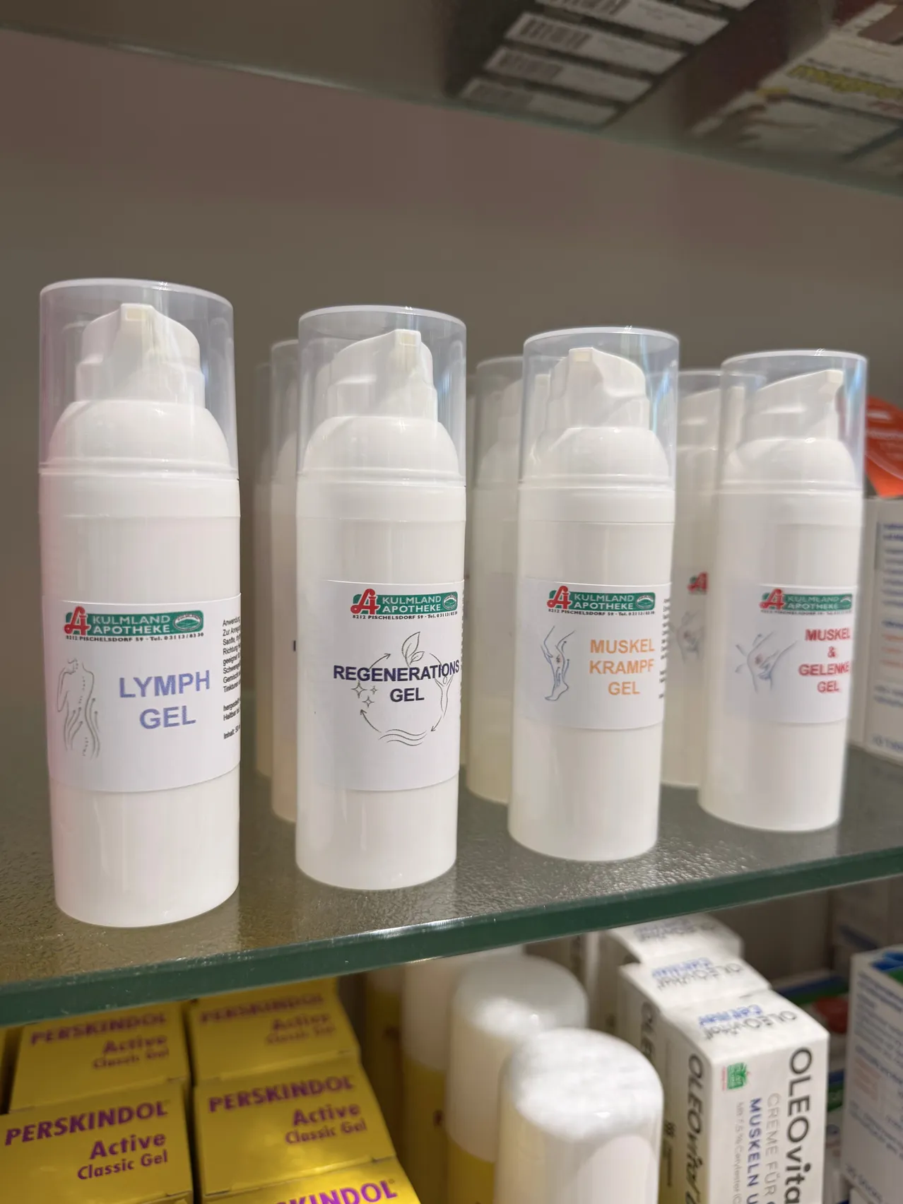 Eine Reihe weißer Flaschen mit durchsichtigen Plastikdeckeln, beschriftet mit 'Lymph Gel', 'Regenerations Gel' und 'Muskel Krampf Gel', ausgestellt auf einem Glasregal in einer Apotheke.