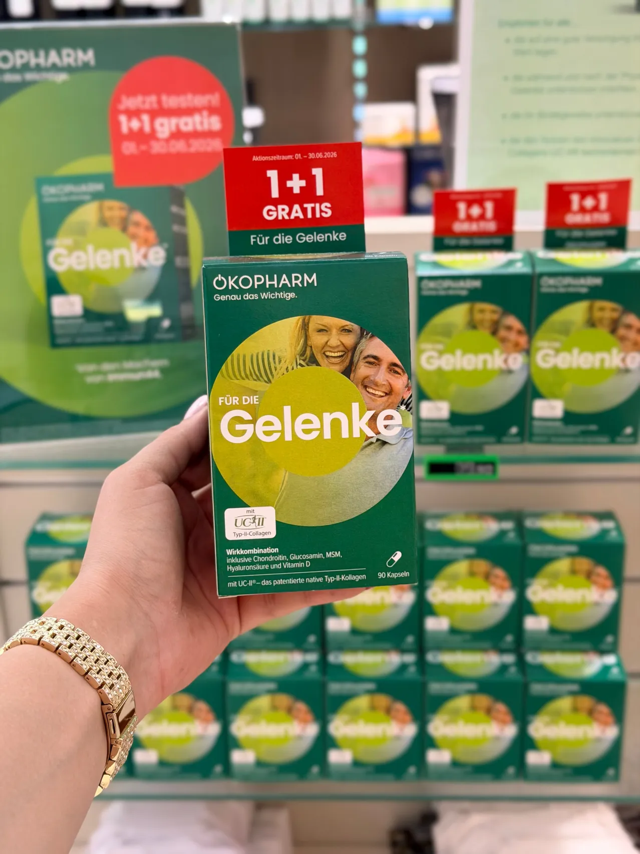 Eine Person hält eine grüne Box des Okopharm Gelenkgesundheitsergänzungsmittels. Es hat einen grünen Deckel mit Bildern eines Mannes und einer Frau. Im Hintergrund befinden sich weitere Boxen von Okopharm.