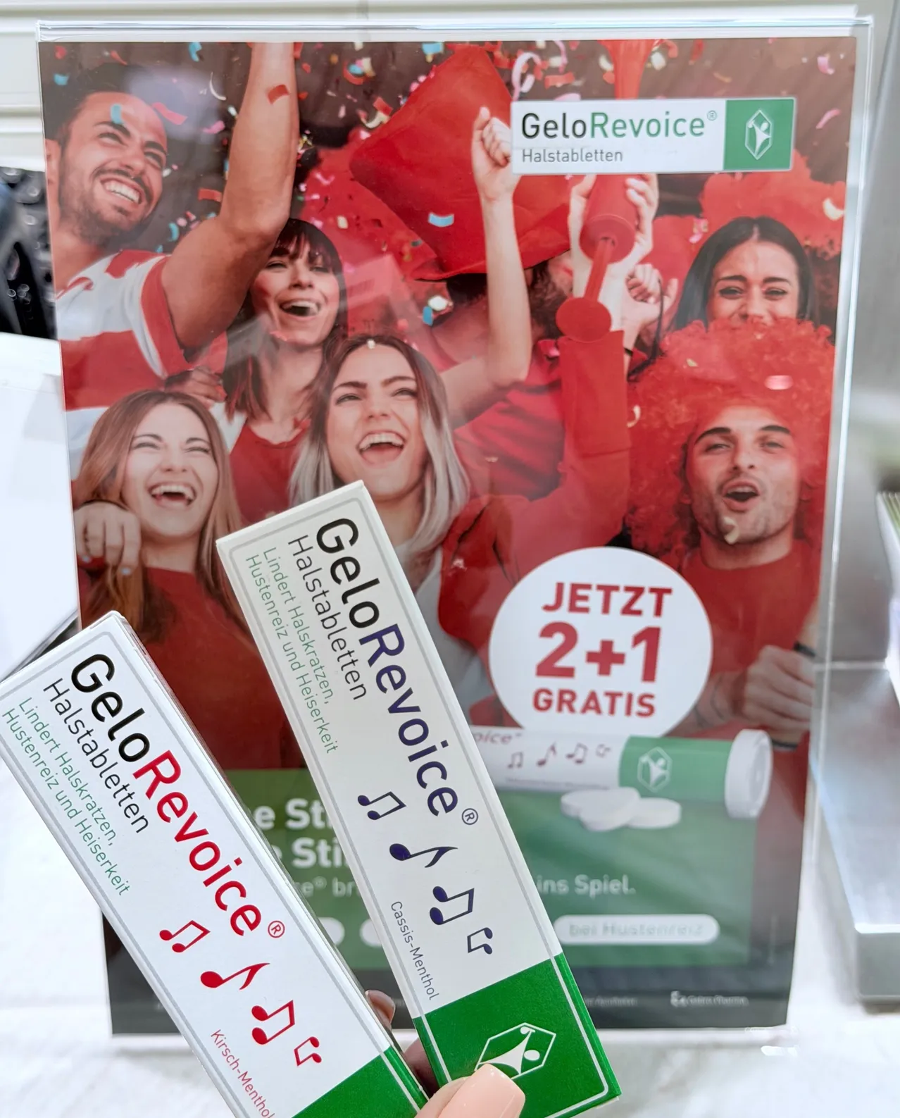 Zwei Packungen GeloRevoice, eine Marke für Halspastillen, werden vor einem Poster gehalten, das ein '2+1 gratis' Angebot anbietet. Das Poster zeigt Menschen, die rote Hüte tragen, lächeln und rote Tassen halten.
