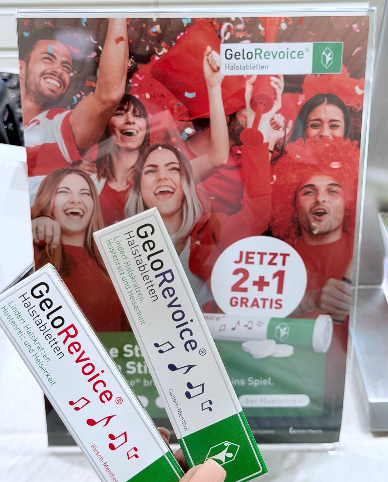 Zwei Packungen GeloRevoice, eine Marke für Halspastillen, werden vor einem Poster gehalten, das ein '2+1 gratis' Angebot anbietet. Das Poster zeigt Menschen, die rote Hüte tragen, lächeln und rote Tassen halten.