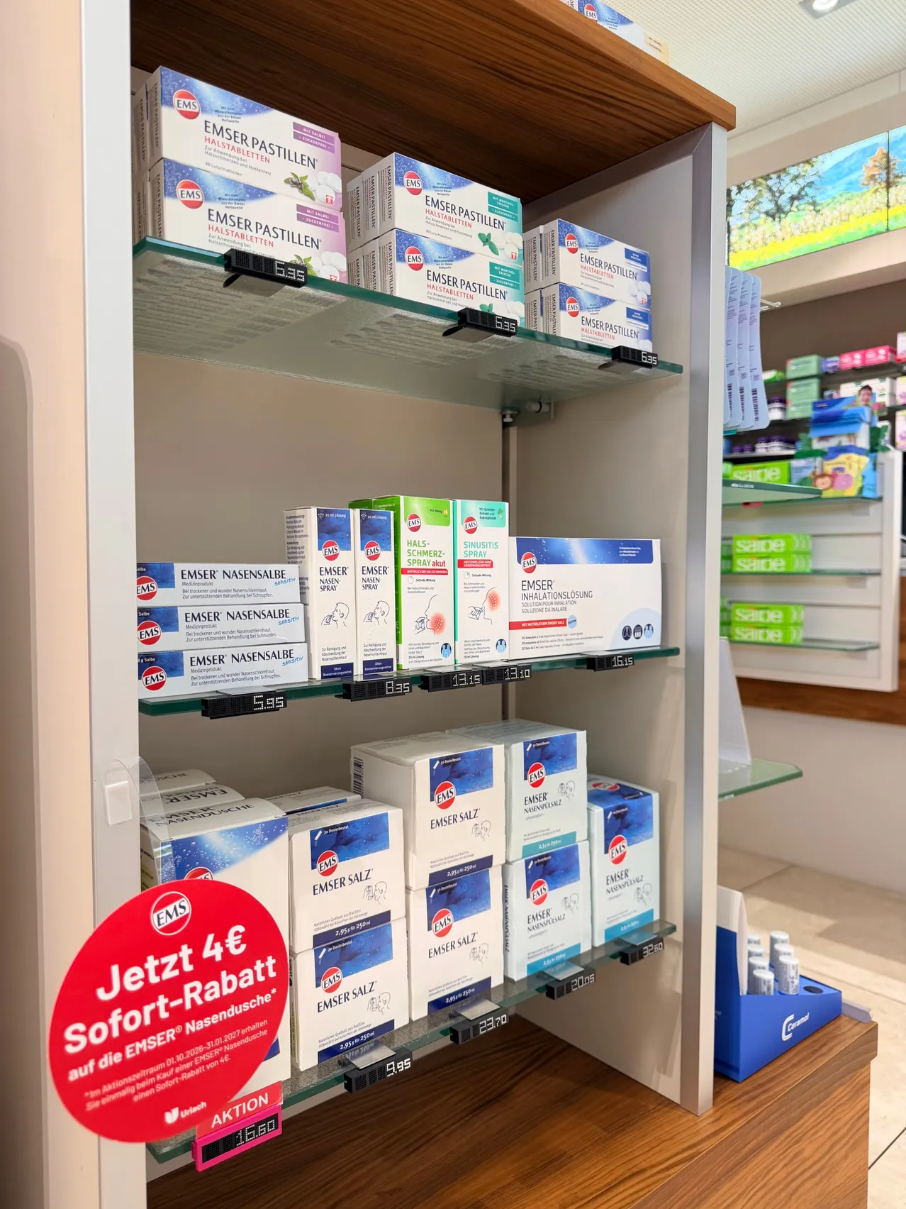 Ein Apothekenregal mit verschiedenen EMSER-Produkten, einschließlich Nasensprays und Pastillen. Die Produkte sind ordentlich auf Glasregalen mit Etiketten angeordnet. Ein rotes Schild mit dem Text 'Jetzt 4€ Sofort-Rabatt' ist sichtbar und zeigt ein Rabattangebot an.