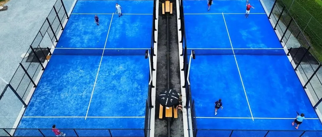 Eine Draufsicht auf mehrere Tennisplätze mit Spielern in Aktion. Spieler in roten und weißen Uniformen sind auf gegenüberliegenden Seiten der Plätze positioniert. Die Plätze sind von Glaswänden umschlossen und haben gelbe Bänke.