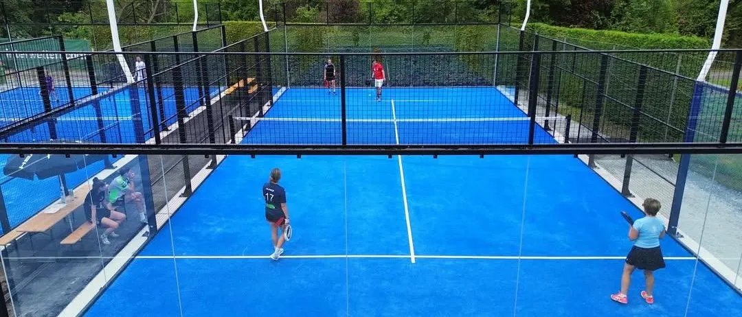 Ein Outdoor-Tennisplatz mit blauer Oberfläche und schwarzen Zäunen. Spieler sind auf dem Platz, und einer hält eine Schläger. Der Platz ist von Grün umgeben.