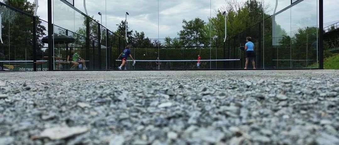 Zwei Frauen spielen Tennis auf einem Platz mit Schotterboden. Eine Spielerin schwingt den Schläger, während eine andere darauf wartet, den Ball zu treffen. Zuschauer sitzen auf Bänken.