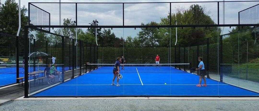 Eine Gruppe von Menschen spielt Tennis auf einem eingezäunten Platz. Eine Person in Rot serviert und eine andere ist dabei, den Ball zu schlagen. Zwei andere beobachten.