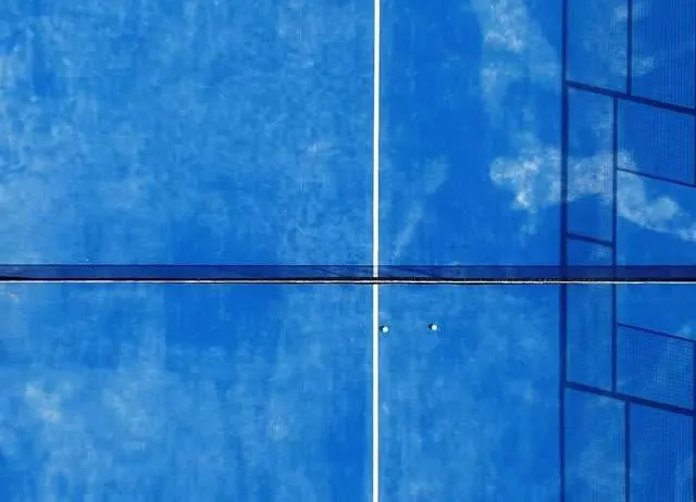 Ein Tennisplatz mit blauen und weißen Oberflächen, der durch ein Netz geteilt ist. Der Platz spiegelt den Himmel wider, und Schatten werden auf die Oberfläche geworfen.