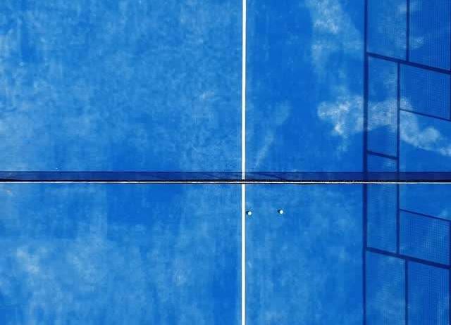 Ein Tennisplatz mit blauen und weißen Oberflächen, der durch ein Netz geteilt ist. Der Platz spiegelt den Himmel wider, und Schatten werden auf die Oberfläche geworfen.