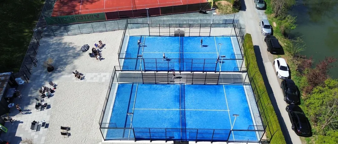 Luftaufnahme von drei blauen Paddelplätzen, die von einem Zaun umgeben sind. Mehrere Personen spielen oder stehen auf den Plätzen. Zuschauer sitzen an den Seitenlinien. Im Hintergrund ein roter Tennisplatz und ein Parkplatz mit geparkten Autos.