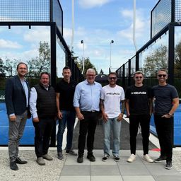 Sechs Männer stehen in der Nähe eines Tennisplatzes. Sie tragen alle eine Sonnenbrille und Freizeitkleidung. Der Hintergrund ist ein blauer Himmel mit Wolken.