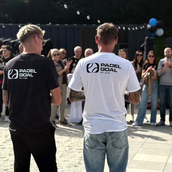 Zwei Männer stehen vor einer Menge, die 'Padel Goal'-Shirts tragen, einer in Schwarz und der andere in Weiß. Hinter ihnen klatschen und bewundern die Menschen die Veranstaltung.