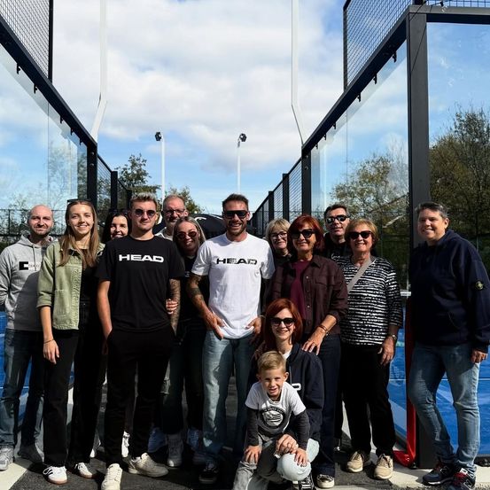 Eine Gruppe von Menschen steht in der Nähe eines Tennisplatzes mit Glasbarrieren. Einige tragen Sonnenbrillen, und ein Kind hält einen Ball. Sie scheinen ein Gruppenfoto zu machen.