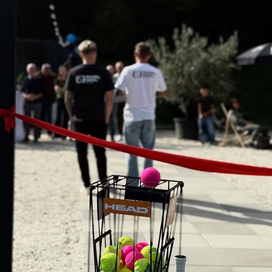 Ein Tennisplatz mit einem Korb voller HEAD Tennisbällen. Zuschauer stehen hinter einer roten Bandbarriere und beobachten das Spiel.