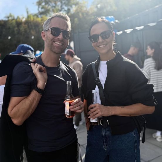 Ein Mann und eine Frau, beide mit Sonnenbrillen, lächeln und stehen zusammen, mit Flaschen in der Hand. Sie sind draußen, und es gibt andere Leute und Bäume im Hintergrund.