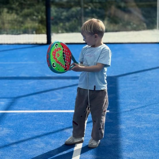 Ein junger Junge mit Brille hält einen bunten Schläger auf einem blauen Tennisplatz. Er sieht nach rechts.