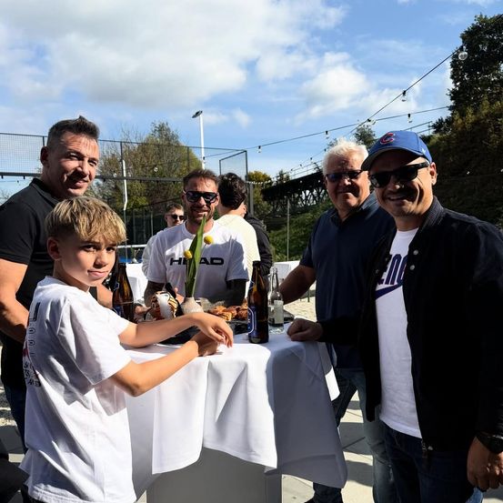 Eine Gruppe von Männern und einem Jungen stehen um einen Tisch mit Getränken und Essen. Der Junge lächelt und hat seine Hand auf dem Tisch. Dahinter ist ein Zaun und eine Brücke.