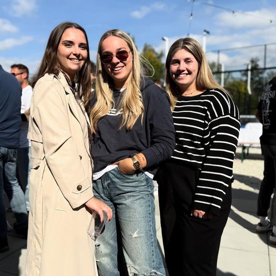 Drei Frauen in Freizeitkleidung stehen in einem Außenbereich, lächeln und posieren für ein Foto. Hinter ihnen steht eine Gruppe von Menschen.