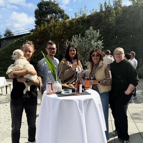 Eine Gruppe von Menschen steht um einen Tisch mit Getränken und einem Hund und lächelt für die Kamera.