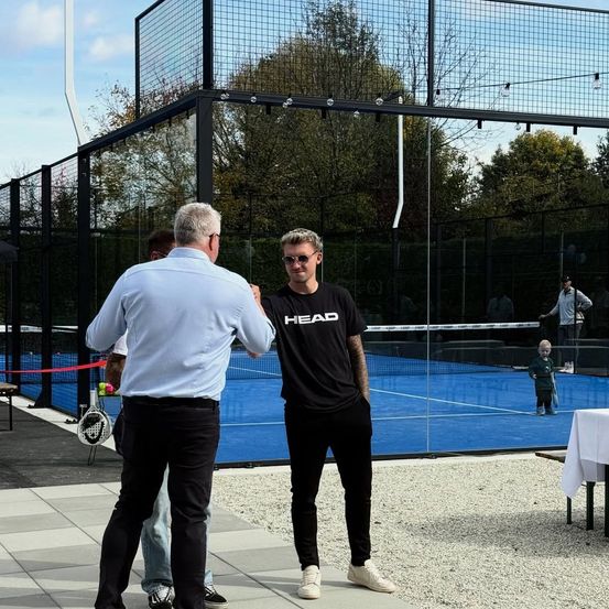 Ein Mann in einem HEAD-Shirt steht neben einem blauen Tennisplatz. Ein Mann in einem weißen Hemd schüttelt ihm die Hand. Ein kleiner Junge spielt im Hintergrund Tennis.