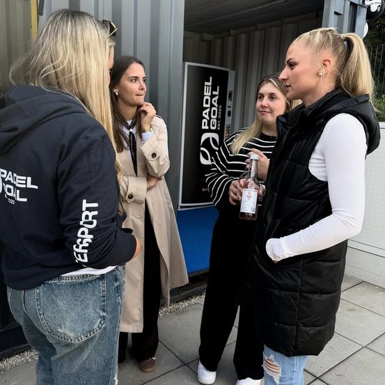 Vier Frauen stehen vor einem Container. Eine hält eine Flasche, eine andere trägt eine Jacke mit dem Wort 'GOAL'. Eine dritte trägt einen Mantel und die vierte trägt eine Kapuzenjacke. Sie scheinen etwas zu besprechen.