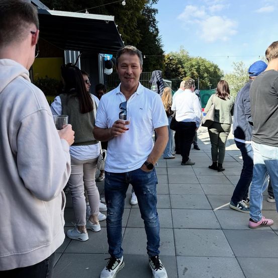 Ein Mann in einem weißen Hemd und Jeans steht unter einer Menschenmenge auf einem Bürgersteig. Er hält ein Getränk in der Hand.