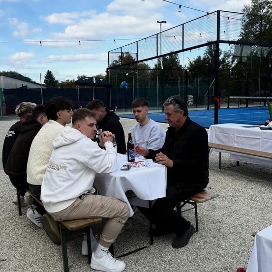 Eine Gruppe von Männern sitzt auf einer Bank um einen Tisch herum, möglicherweise bei einer Besprechung. Sie befinden sich in einem Außenbereich mit einem eingezäunten Tennisplatz im Hintergrund.