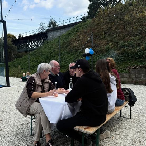 Eine Gruppe von Menschen sitzt draußen um einen Tisch mit Luftballons und einer Brücke im Hintergrund.