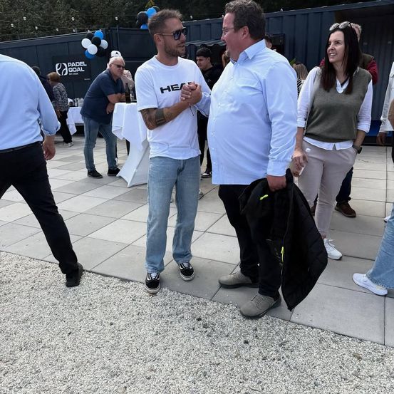Eine Gruppe von Menschen steht in einem Außenbereich. Zwei Männer unterhalten sich, einer in einem weißen Shirt, der andere in Hellblau. Hinter ihnen lächelt eine Frau. In der Nähe sind Tische und Luftballons aufgestellt.