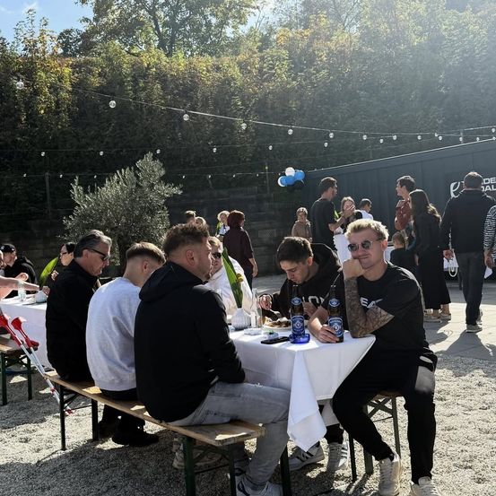 Eine Gruppe von Menschen in Freizeitkleidung sitzt um einen Tisch in einer Outdoor-Umgebung, einige stehen und unterhalten sich. Auf dem Tisch sind Flaschen und Teller, und dekorative Lichter hängen darüber.