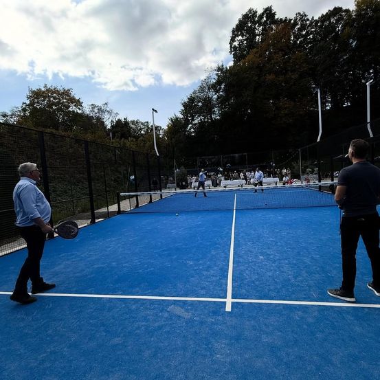 Zwei Männer spielen Tennis auf einem blauen Platz, einer hält eine Racket. Ein Schiedsrichter steht an der Seitenlinie. Zuschauer sitzen im Hintergrund.