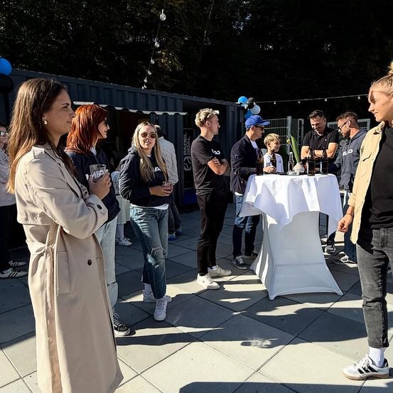 Mehrere Personen, darunter eine Frau in einem beigen Mantel, versammeln sich um einen Tisch mit Flaschen und Bechern, möglicherweise bei einer gesellschaftlichen Veranstaltung.