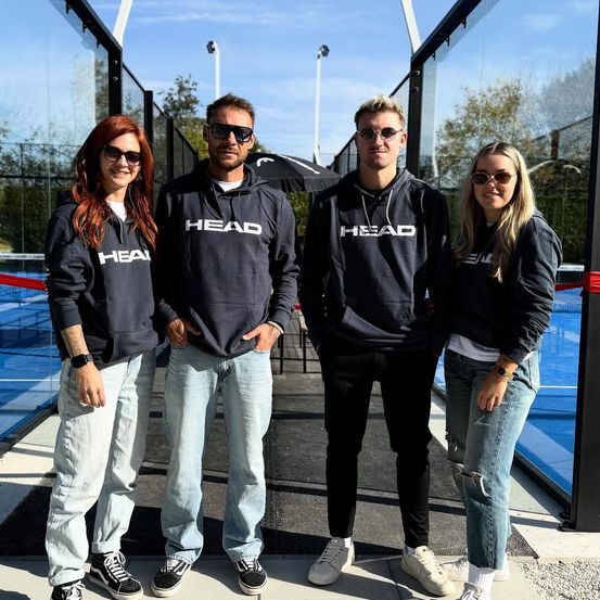 Vier Menschen in schwarzen HEAD-Sweatshirts und mit Sonnenbrillen stehen vor einem Tennisplatz. Ein schwarzer Regenschirm befindet sich hinter ihnen.