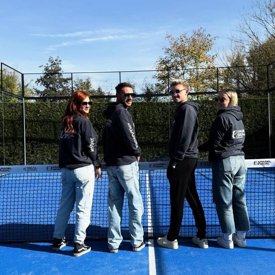 Vier Personen in dunklen Kapuzenjacken und Jeans stehen zusammen auf einem blauen Tennisplatz, mit einem Zaun und Bäumen im Hintergrund.