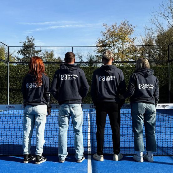 Vier Personen stehen auf einem blauen Padel-Court und blicken auf das Netz. Sie tragen passende Hoodies mit dem Logo 'Padel Goal' auf der Vorderseite. Der Himmel ist blau mit einigen Wolken, und Bäume mit Herbstlaub sind im Hintergrund zu sehen.