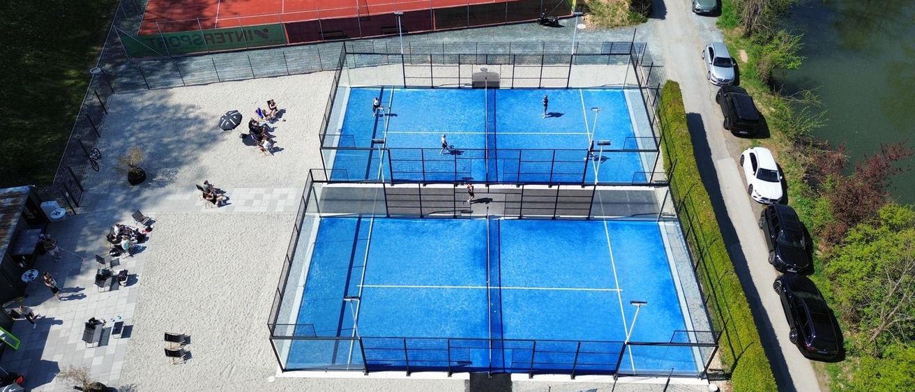 Eine Vogelperspektive eines Tenniskomplexes mit drei blauen Plätzen und einem roten Platz, mehrere Personen spielen, andere Zuschauer an den Seiten und Autos auf der Straße geparkt.