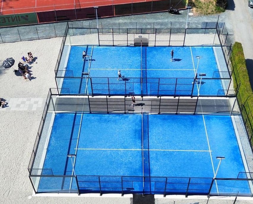 Eine Vogelperspektive eines Tenniskomplexes mit drei blauen Plätzen und einem roten Platz, mehrere Personen spielen, andere Zuschauer an den Seiten und Autos auf der Straße geparkt.