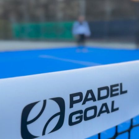 Ein verschwommener Blick auf eine Person auf einem blauen Padel-Court mit einem weißen Netz. Das Netz hat das Logo 'Padel Goal'.