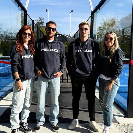 Vier Personen stehen nebeneinander in HEAD-Kapuzenpullovern. Sie sind draußen vor einer Glaswand und einem Tennisplatz.