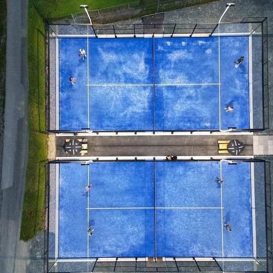 Eine Vogelperspektive von zwei umzäunten blauen Tennisplätzen mit Spielern. Ein Platz hat zwei Spieler in der Mitte und der andere hat mehrere Spieler. Bänke und Straßenlaternen sind sichtbar.