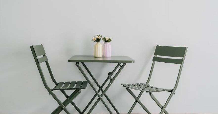 Zwei grüne Stühle umgeben einen kleinen grünen Tisch mit zwei Vasen darauf. Die Vasen haben weiße und gelbe Blumen. Der Tisch und die Stühle stehen auf einem Holzboden vor einer weißen Wand.