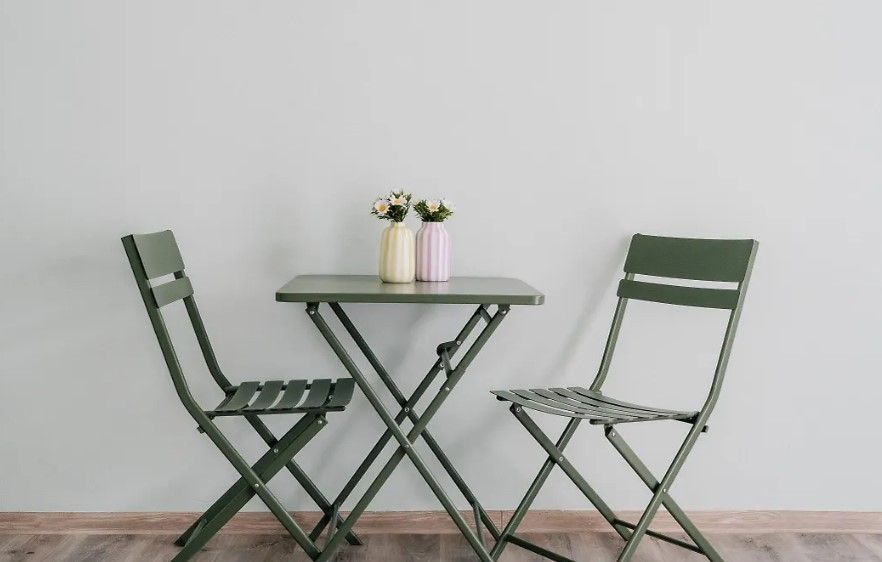 Zwei grüne Stühle umgeben einen kleinen grünen Tisch mit zwei Vasen darauf. Die Vasen haben weiße und gelbe Blumen. Der Tisch und die Stühle stehen auf einem Holzboden vor einer weißen Wand.