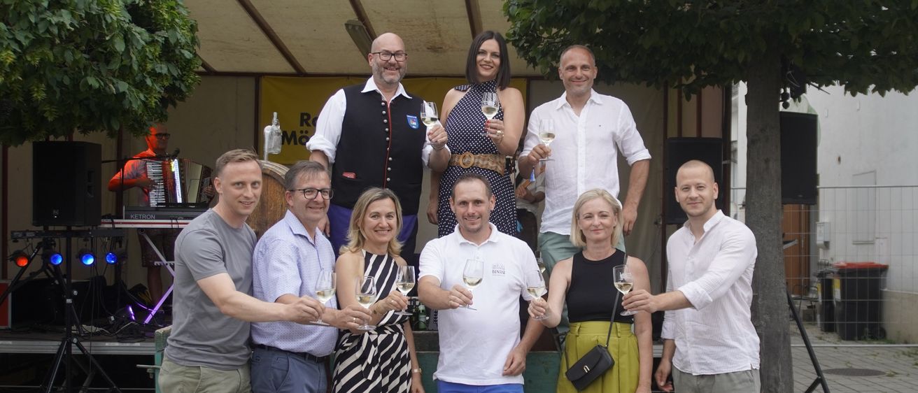 Eine Gruppe von Menschen steht zusammen, lächelt und hält Weingläser in der Hand. Sie sind lässig gekleidet, einige tragen eine Brille und Turnschuhe. Eine Frau in einem Punktekleid und ein Mann in einem Vest stehen hinten. Sie scheinen sich auf einer Weinprobe zu befinden.