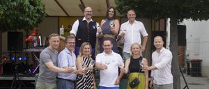 Eine Gruppe von Menschen steht zusammen, lächelt und hält Weingläser in der Hand. Sie sind lässig gekleidet, einige tragen eine Brille und Turnschuhe. Eine Frau in einem Punktekleid und ein Mann in einem Vest stehen hinten. Sie scheinen sich auf einer Weinprobe zu befinden.