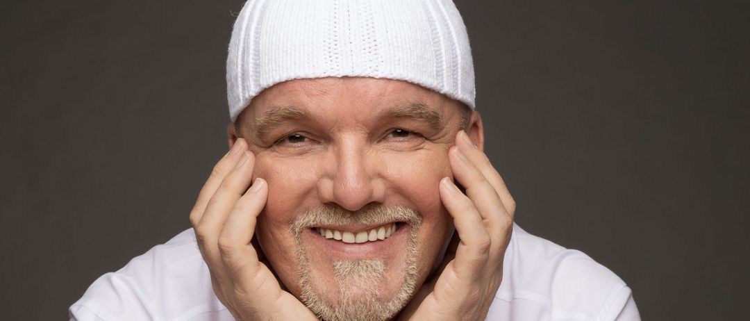 Ein Mann mit weißer Mütze und weißem Hemd posiert lächelnd mit den Händen an den Wangen, sitzend an einem Holztisch.