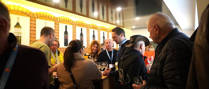 Eine Gruppe von Menschen steht in einer Weinkellerei um eine Bar herum, unterhält sich und trinkt Wein. Die Bar ist mit Flaschen und einer gelben Wand dekoriert.