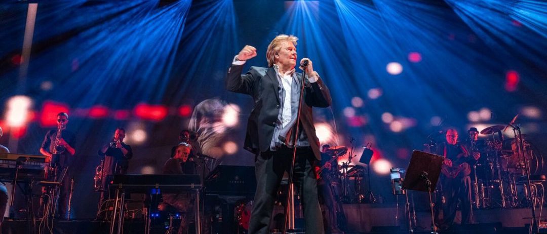 Ein Mann steht mit einem Mikrofon auf der Bühne und tritt vor einer Band auf. Die Bühne ist mit blauen und roten Lichtern beleuchtet. Andere Musiker sitzen hinter ihm und spielen verschiedene Instrumente.